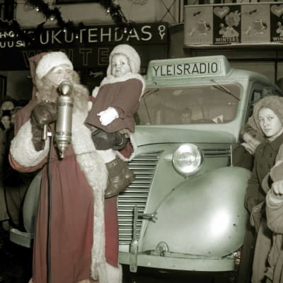 Joulupukki, jolla on sylissään pieni tyttö. Heidän takanaan on Yleisradion auto. Valokuvaa sävytetty Elävän arkiston toimituksessa.