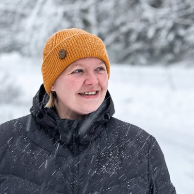 Nainen seisoo lumisateessa ja katsoo sivuun hymyillen.