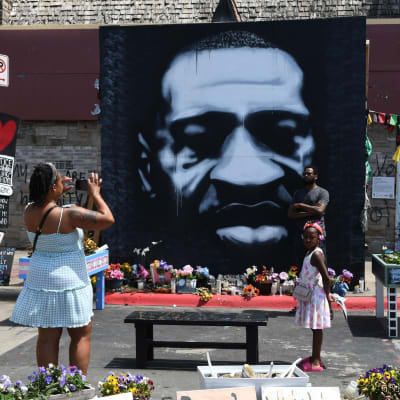 Platsen där George Floyd dog i Minneapolis. Vid en stor väggmålning av Floyd står en svart man medan en svart kvinna tar en bild av honom. En svart flicka står i närheten och tittar på alla blomsterhyllningar på platsen.