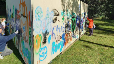 En triangelformad vägg eller ett plank som placerats ute på en gräsmatta. Barn och ungdomar målar på det.