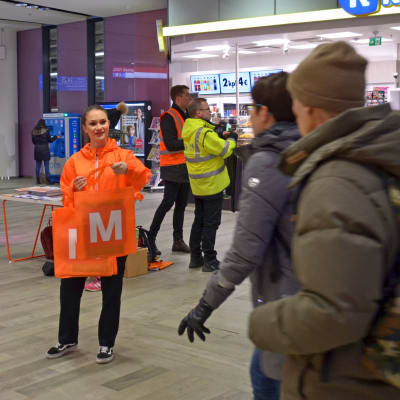 Orangeklädd personal delar ut metrokassar till passagerare i Mattby bussterminal.