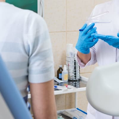 En läkare drar på sig blå plasthandskar medan en kvinna som man endast ser ryggen av sitter i en gynekologisk undersökningsstol.