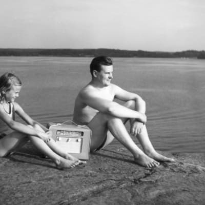 Mies ja lapsi kuuntelevat radiota rantakalliolla, mustavalkoisessa valokuvassa.
