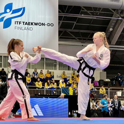 Ariella Figueroa ja Mira Sjövall kohtaavat toisensa taekwondo-ottelussa. Oikealla oleva nainen on nostanut oikean jalkansa potkuun, vasemmalla oleva nainen torjuu sen. 