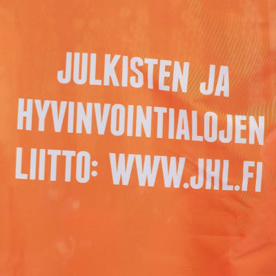 Oranssinen lippu, jossa valkoisella teksti JULKISTEN JA HYVINVOINTIALOJEN LIITTI: WWW.JUHL.FI. Taustalla ihmisiä.
