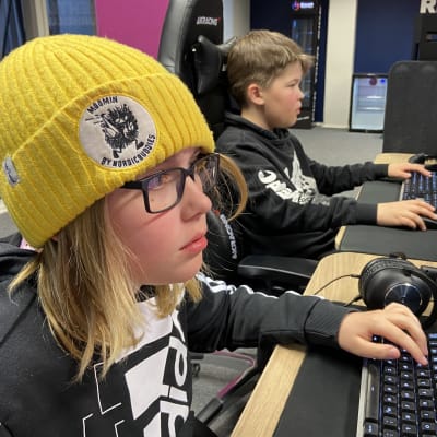 Kaksi alle pikkupoikaa istuu vierekkäin tietokoneiden äärellä pelaamassa tietokonepelejä. Etummaisella on keltainen pipo päässä ja silmälasit