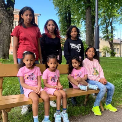 Sju flickor i olika åldrar står uppradade för bild vid en bänk i en park.