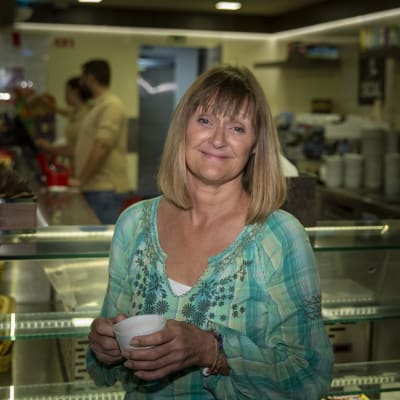 En medelålders kvinna med blont går och blå tröja står i en butik. I händerna håller hon en kaffekopp.