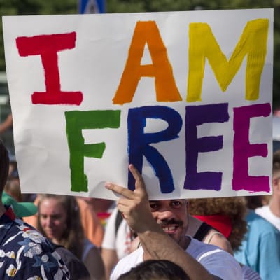Demonstranter håller upp en skyld med texten "I AM FREE" i regnbågens färger.
