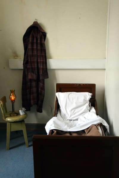 En säng, en gammal stol med oljelampa på, en morgonrock hänger på väggen.