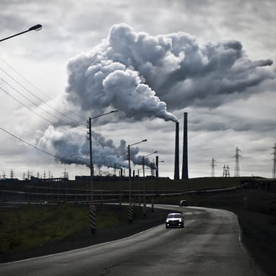 En ensam bil på en väg i ett grått landskap som präglas av elstolpar. Centralt syns flera fabriksskorstenar som spyr ut rök.