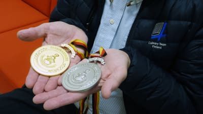 En man håller i två medaljer. Den ena är i guld och den andra i silver. På jackan står det Culinary Team Finland.
