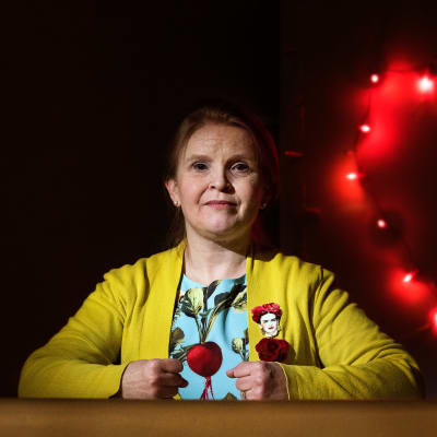 Jenni-Juulia Wallinheimo-Heimonen istuu pöydän takana kädet nyrkissä pöydän päällä. Tummassa taustassa vasemmalla puolella valonauhasta muodostuva sydän.