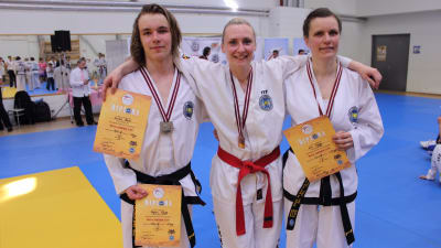Tre personer i vita taekwondodräkter, medaljer hänger runt halsen och de har diplom i handen.