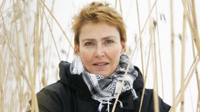 Författaren Minna Lindeberg iklädd en svart jacka och svartvit sjal står ibland vass och tittar in i kameran.