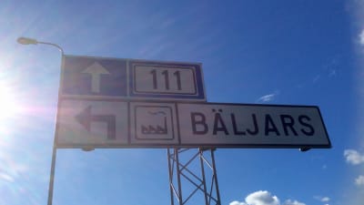 En skylt som visar att Bäljars industriområde (i Karis) är till vänster. Solen skiner, himlen är sommarblå med vita moln.