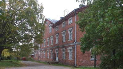 En rödtegelbyggnad i 2-3 våningar, en gammal arbetarkasern i Billnäs bruk. Gräsplan, grusgång, lövträd.