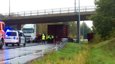 Efter en trafikolycka på en regnig väg med en bro som går över. Poliser och räddningsmanskap i gula västar på plats. Containrar ligger på vägen under bron.