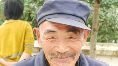 En gammal man i blågrå keps och blågrå skjorta. Han har grått skägg och mustach och han ser glad ut.
