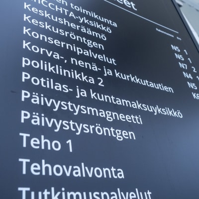 Musta sairaalan poliklinikkojen ja palvelupisteiden opastuskyltti, jossa valkoisella tekstillä eri osastojen nimiä ja lyhenteitä.