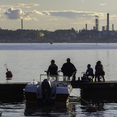 Perhe istuu laiturilla kalastamassa Porvoon öljyjalostamon siintäen taivaanrannassa.