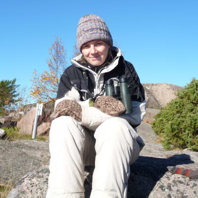 Kvinna, Solja Kvarnström, sitter på berg med mössa på huvudet och kikare runt halsen. Soligt höstväder.