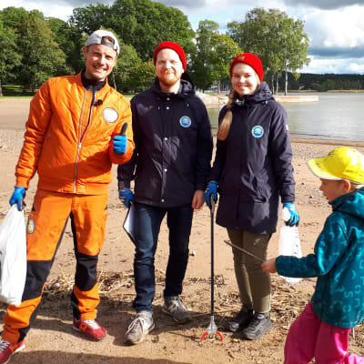 Programledaren Jontti Granbacka står på Runsala strand tillsammans med representanter för Håll Skärgården Ren rfs projekt Snygg beach, på bilden också ett skräpplockande barn