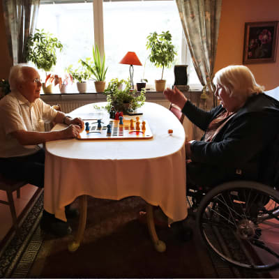 En äldre man sitter och spelar spel med en äldre kvinna som sitter i rullstol.