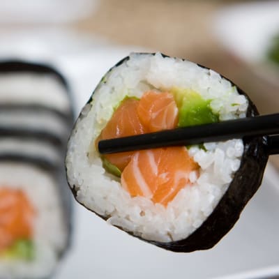 En bit rullad maki-sushi mellan två matpinnar med flera sushibitar i bakgrunden.
