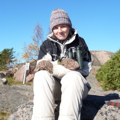 Kvinna, Solja Kvarnström, sitter på berg med mössa på huvudet och kikare runt halsen. Soligt höstväder.