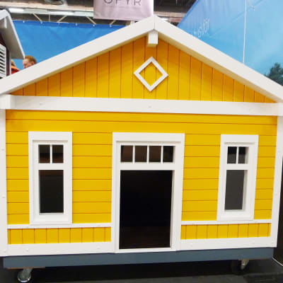 En gul liten byggand (bod) med vita knutar. Står tätt intill ett rött hus.