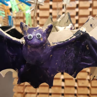 Lapsen askartelema ja violetiksi maalaama lentävä lepakko, jolla on muovisilmät. Halloween-koristeeksi tarkoitettuja maalattuja lepakoita on kuivumassa työpajassa pyykkipojilla pieneen telineeseen kiinnitettyinä.