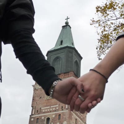 Två kvinnohänder med förlovningsringar håller i varandra med Åbo domkyrka i bakgrunden.