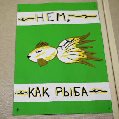 Opetustaulu, jossa piirros kalasta. Tekstissä lukee venäjäksi: mykkä kuin kala. 
