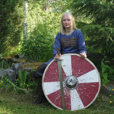 Emelie Lytz sitter på en sten i klädd vikingakläder med ett svärd och ett sköld framför sig.