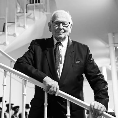 Modeskaparen Pierre Cardin poserar vid en vit trappa i svart kostym. Bilden är svartvit.