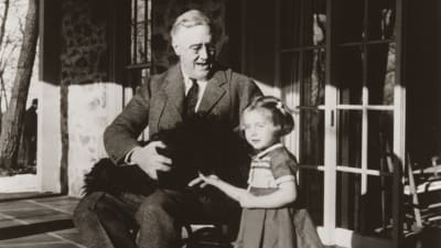 Franlin D. Roosevelt som sitter i en rullstol. I famnen har han en hund och bredvid honom står ett barn, en flicka.
