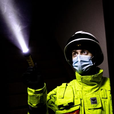 En brandmästare i gul jacka, vit hjälm och munskydd lyser med sin ficklampa i ett sotigt utbränt rum.