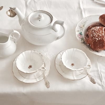 Ett dukat bord med vit bordduk, vit tekanna och vita koppar. Ett blommigt fat med bullar.