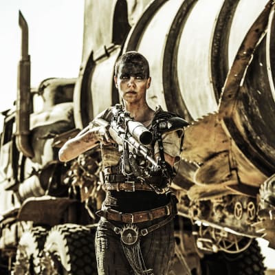 Furiosa (Charlize Theron) kommer gående mot kameran med ett stort vapen i händerna. 