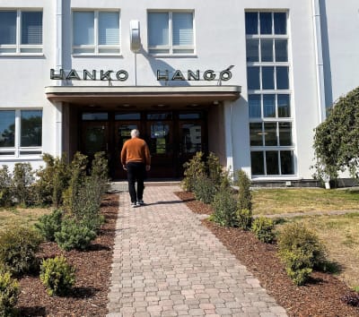 En vit stationsbyggnad där det står Hangö och Hanko vid ingången. Framför en stenbelagd gång och odlingar. En man vandrar mot ingången.
