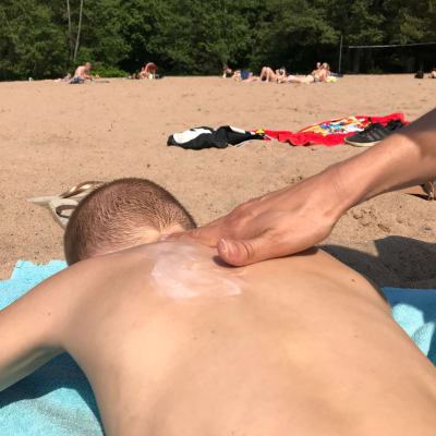Käsi levittää aurinkorasvaa lapsen selkään uimarannalla.