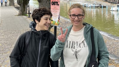 En person med kort, lockigt, mörkt hår och en svart jacka står bredvid en person med glasögon, ljust bakåtkammat hår, en Nirvana-skjorta och grön munkjacka. Den andra personen visar V-tecknet med pekfingret och långfingret. I bakgrunden syns åstranden och Aura å.