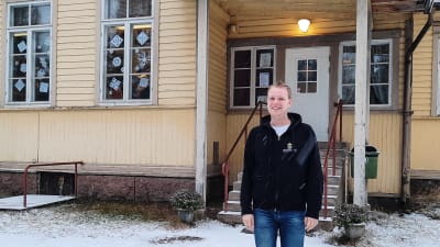 En man i munkjacka står ute i snöregnet framför ett gult trähus.