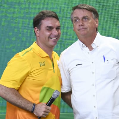 Två män står bredvid varandra: till höger Jair Bolsonaro i kortärmad vit skjorta, till vänster sonen Flávio i kortärmad gul skjorta.