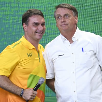 Två män står bredvid varandra: till höger Jair Bolsonaro i kortärmad vit skjorta, till vänster sonen Flávio i kortärmad gul skjorta.
