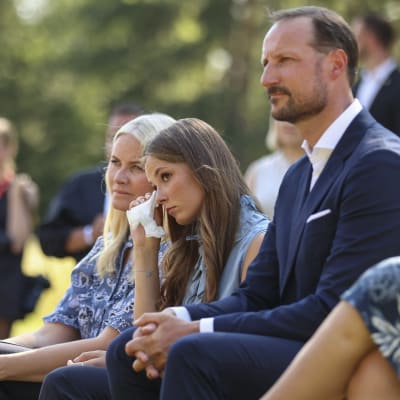 Konrprinsessan Mette-Marit, prinsessan Ingrid Alexandra och kronprins Haakon sitter på bänkar invid Utoyascenen. Ingrid Alexandra torkar bort en tår med en näsduk.