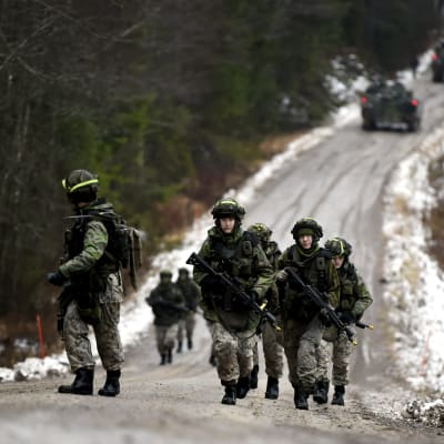 Beväringar deltar i en krigsövning i Kouvola. De bär på vapen och går upp för en backe på en landsväg med skog på båda sidorna.