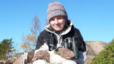 Kvinna, Solja Kvarnström, sitter på berg med mössa på huvudet och kikare runt halsen. Soligt höstväder.