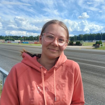 Vaaleaan oranssinpunaiseen huppaariin pukeutunut silmälasipäinen nuori nainen katsoo kameraan ja hymyilee vähän. Taustalla Joensuun ravirata.
